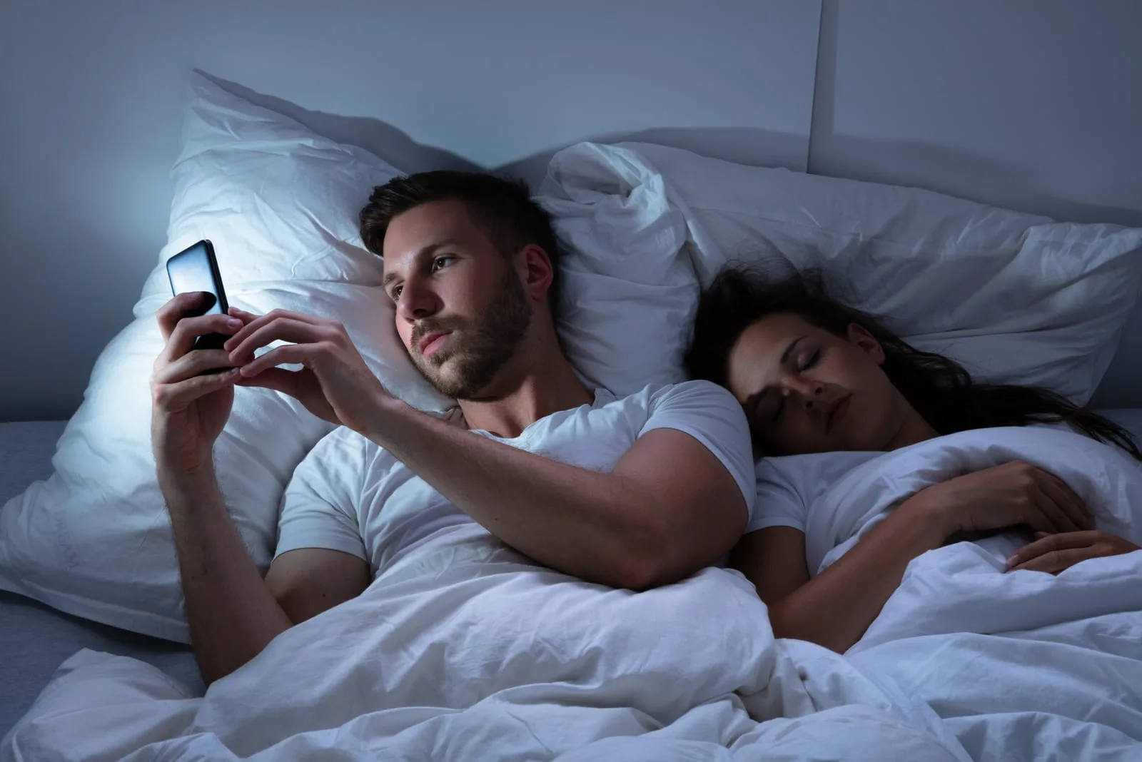 man typing in bed while woman sleeping