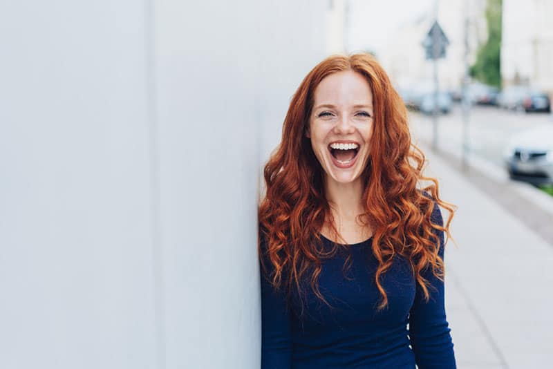 Cute young woman with a lovely sense of humour standing leaning against a white exterior wall with copy space in an urban street laughing at the camera