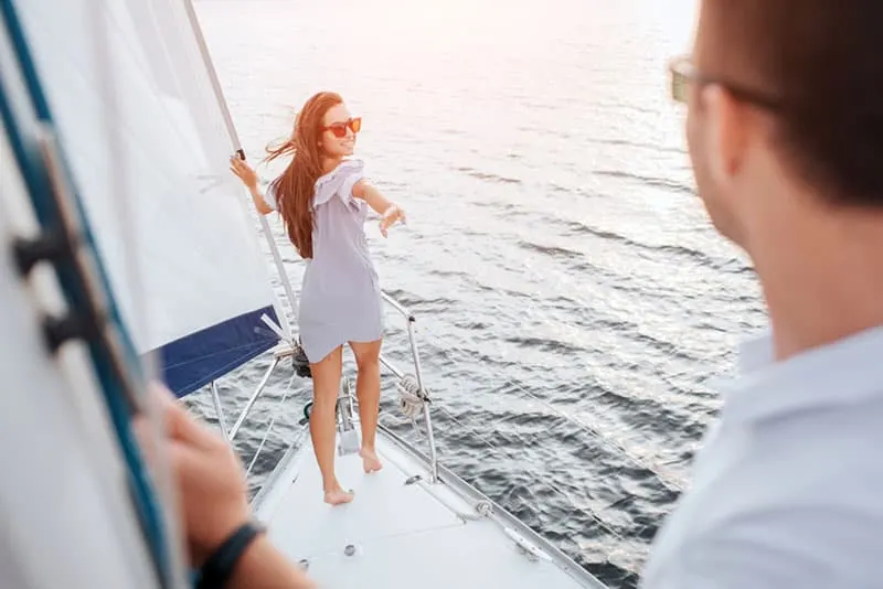 Beutiful and well-built model stands on bow of yacht and holds on tube. She looks behind herself at boyfriend. Brunette try to reaches him with hand and smiles. Young man stands and looks.