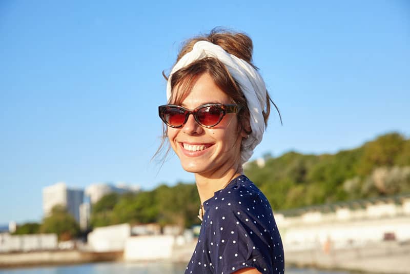 Close-up of happy beautiful dark haired female in headband and sunglasses looking to camera cheerfully, posing outdoor on warm sunny day