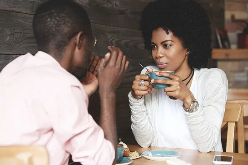 Black girl with Afro hairstyle holding a cup of coffee, listening and looking at her boyfriend with amorous expression. Loving couple enjoying time together at a restaurant on Saint Valentine's Day