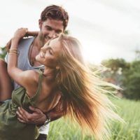 Fotografia de uma jovem mulher a ser carregada pelo namorado num campo relvado. Casal a divertir-se nas suas férias de verão.