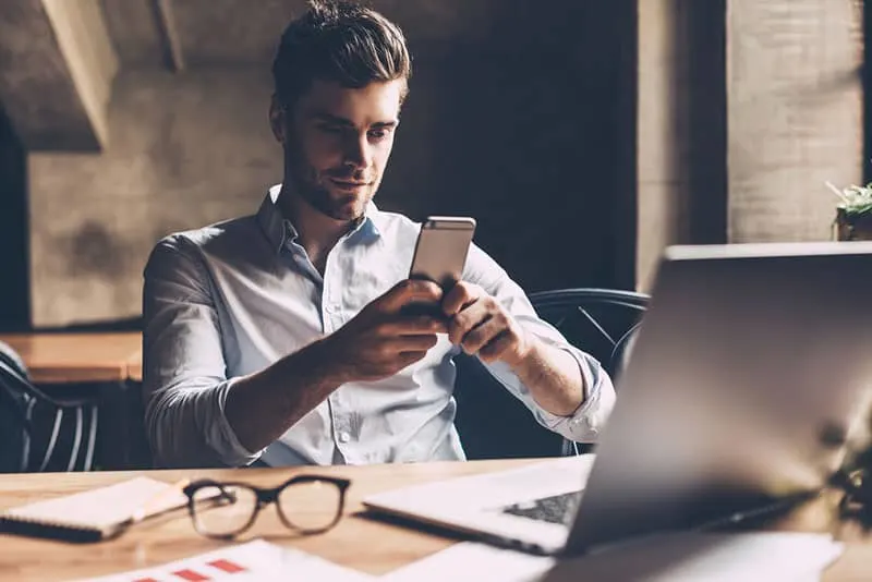 Texting to colleague. Confident young man in smart casual wear holding smart phone and looking at it while sitting at his working place in office