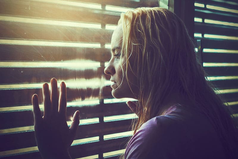 Donna bionda triste e solitaria con i capelli bagnati che guarda attraverso le tende della finestra verso la luce del sole