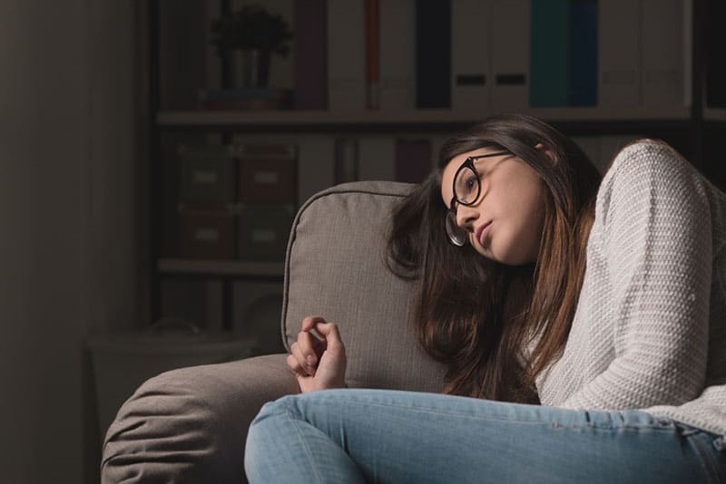Giovane donna triste con gli occhiali seduta sul divano di casa, depressa e sola