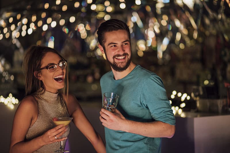 Una pareja de jóvenes adultos se reúnen en un club nocturno para tomar un cóctel. Se ríen y miran a alguien fuera del encuadre.