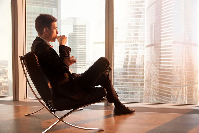 Side view of handsome businessman sitting in comfortable office chair enjoying cup of coffee in the morning, looking through big window at dawn sunrise city, getting motivation for new working day