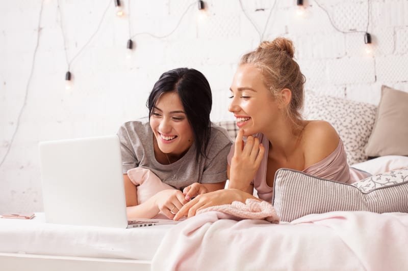 two female friends lying on bed and looking at laptop