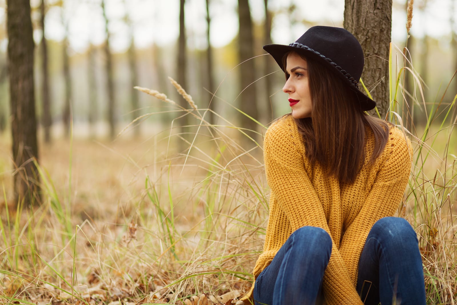a woman in a yellow sweater and black hat sits in the woods on the grass