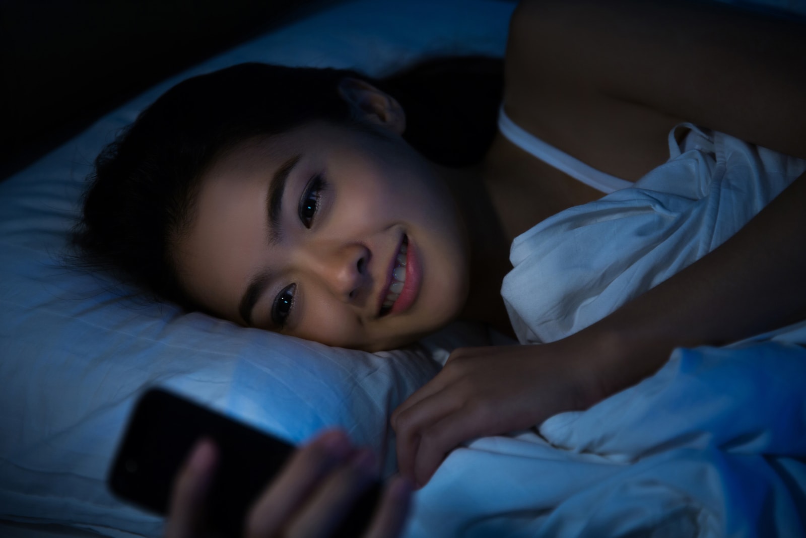 a happy asian woman lies in bed at night and uses a smartphone