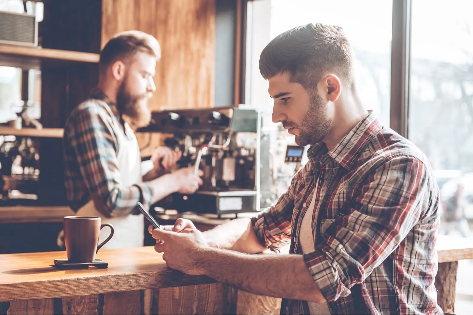 a man in a plaid shirt at the bar drinks coffee and uses a smartphone