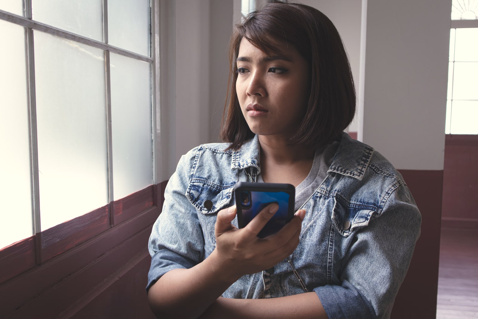 a sad Asian woman is sitting by the window with a smartphone in her hands