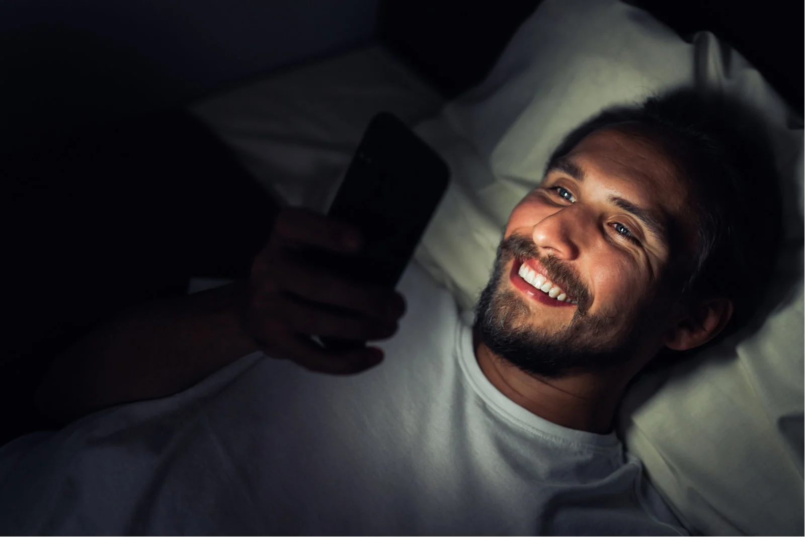 a smiling bearded man lies and reads a message on his smartphone