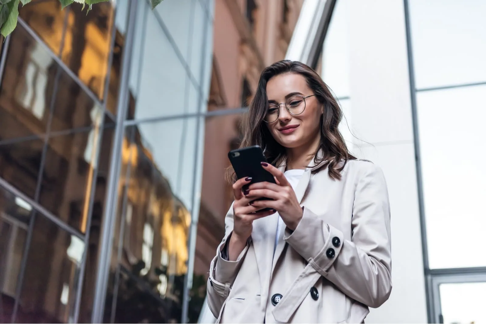 a smiling girl in a white coat stands outside and uses a smartphone