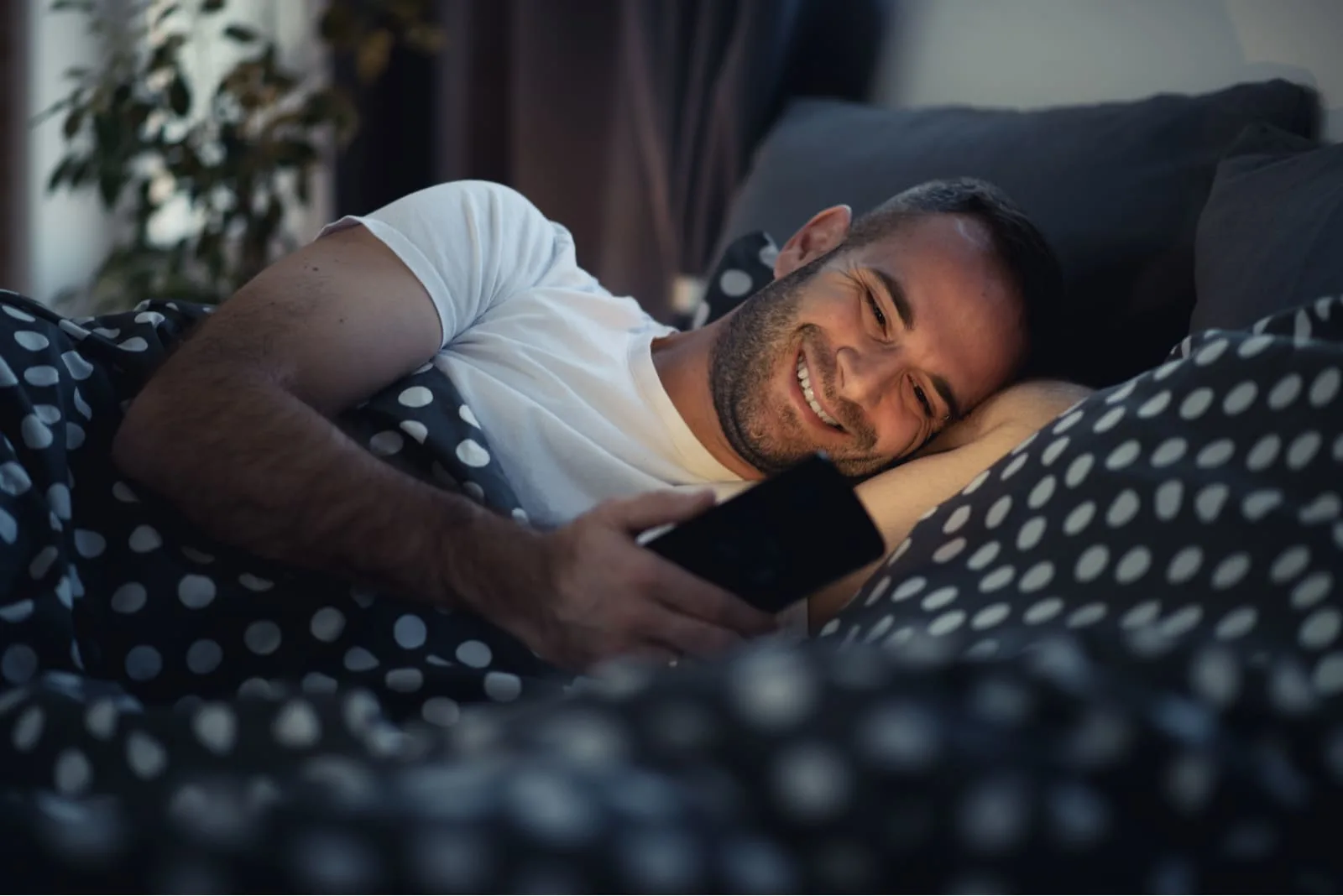 a smiling happy man lying in bed and using a smartphone