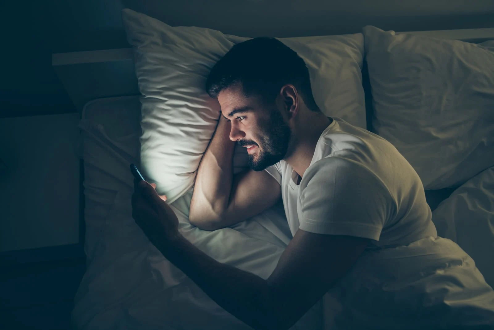 a smiling man in a white T-shirt in the evening lies in bed and watches something on his smartphone