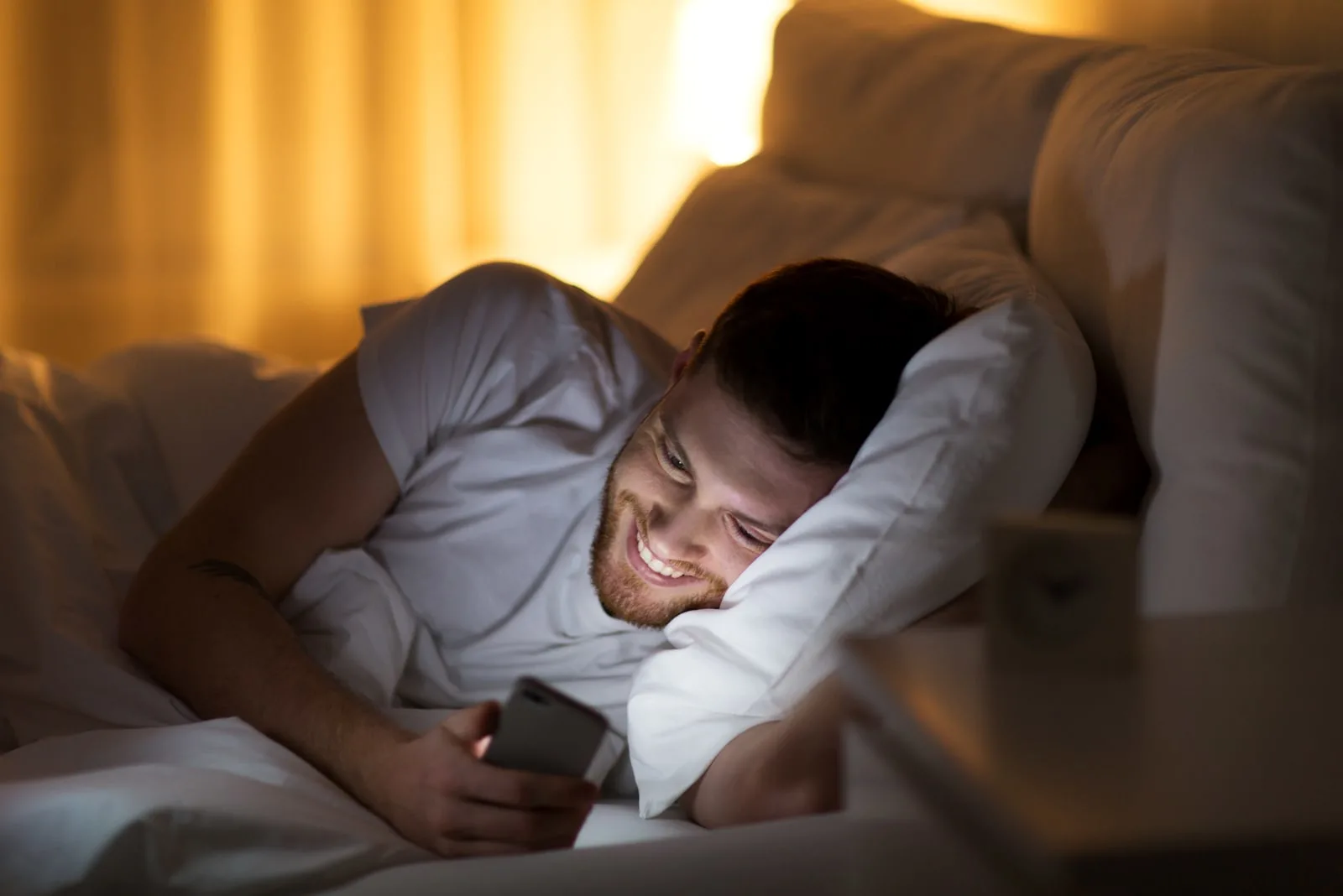 a smiling man lies in the bedroom and uses a cell phone