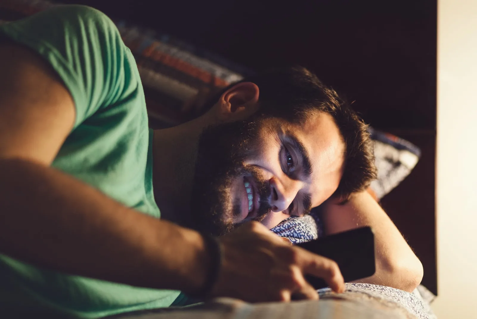 a smiling man with a beard in a green T-shirt lying in bed at night and texting
