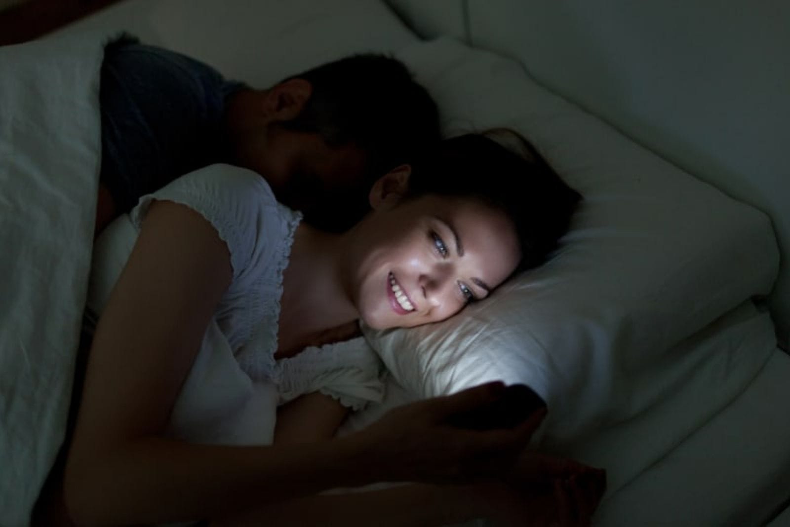 una mujer sonriente en la cama con un hombre dormido utiliza un teléfono móvil