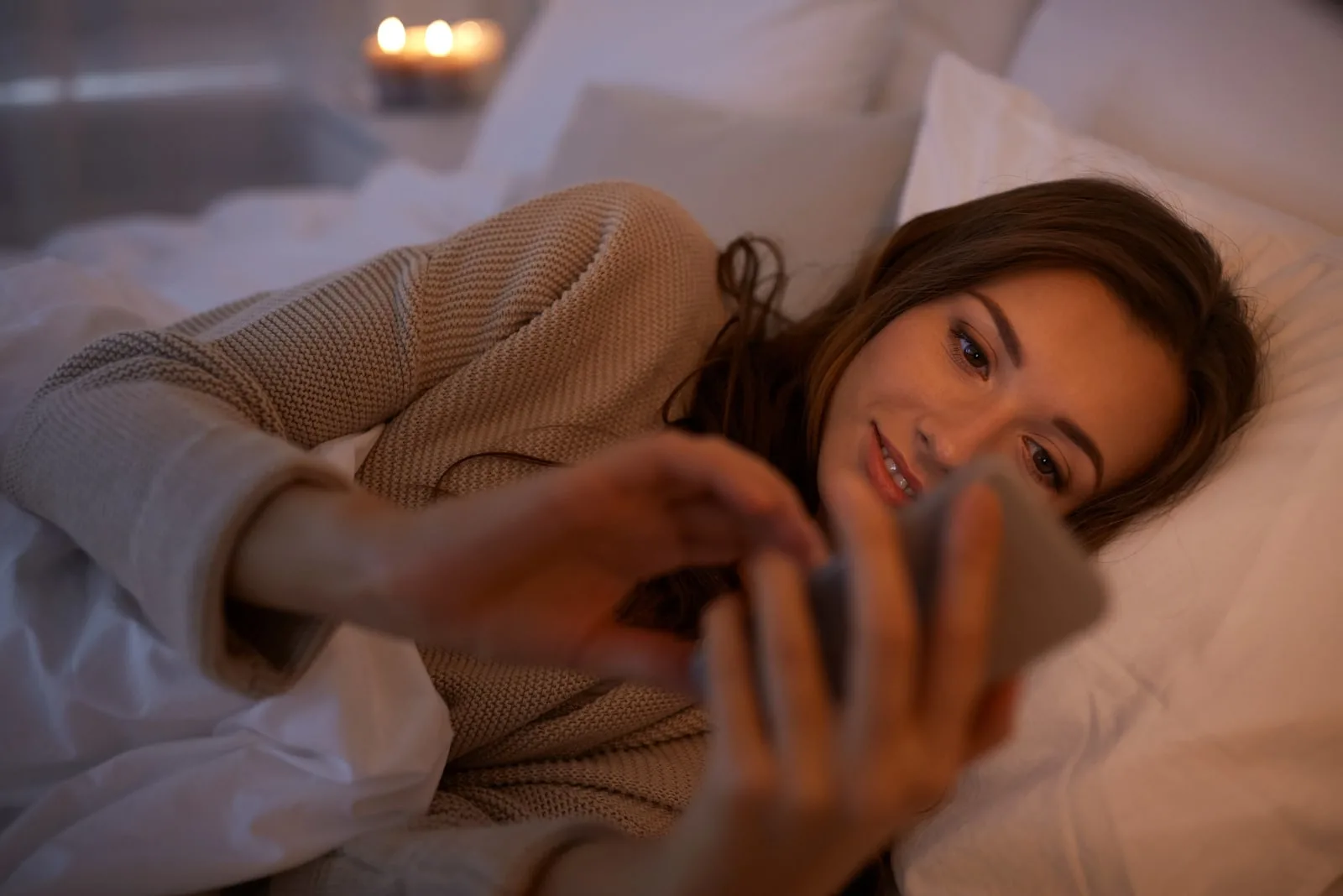 a smiling woman lies in bed and uses a smartphone