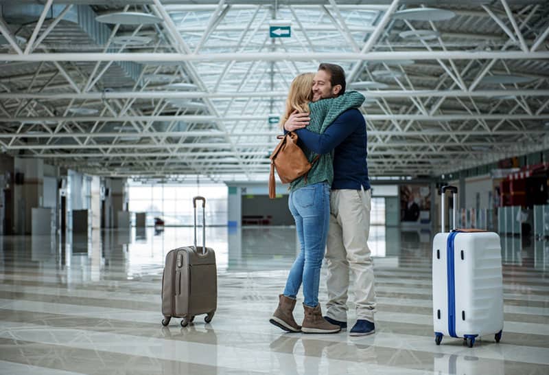 Dois adultos alegres abraçam-se antes da partida, em pé no terminal. As malas estão separadas deles