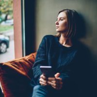 Mujer joven pensativa vestida con traje casual negro mirando por la ventana descansando en un cómodo espacio de coworking. Chica hipster reflexiva con smartphone en manos pensando en el futuro.