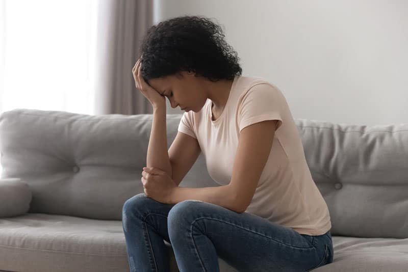 Depressed african American young woman sit on couch at home feel desperate heartbroken suffering from divorce, stressed upset black female thinking having life problems, miscarriage abortion concept