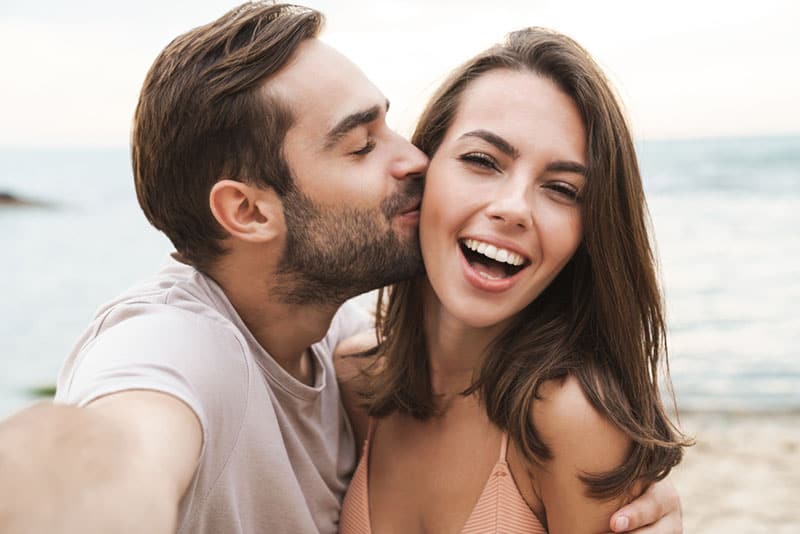 Immagine di un giovane uomo felice che bacia e abbraccia una bella donna mentre si scatta una foto selfie su una spiaggia soleggiata