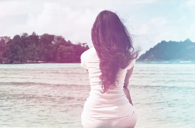 Lonely Woman in White Casual Outfit with Long Black Hair Sitting at Beachfront on a Windy Sunny Day.
