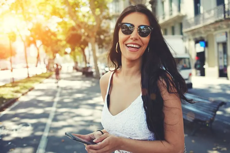 Happy Latin woman in summer sun glasses smiling brightly while holding smart phone strolling outdoors in the city at sunny day,female person using mobile standing with composition copy space on a side