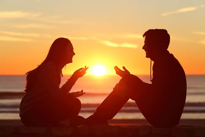 Vista laterale di un corpo intero di due amici o silhouette di coppia di adolescenti seduti a parlare all'alba sulla spiaggia con il sole al centro