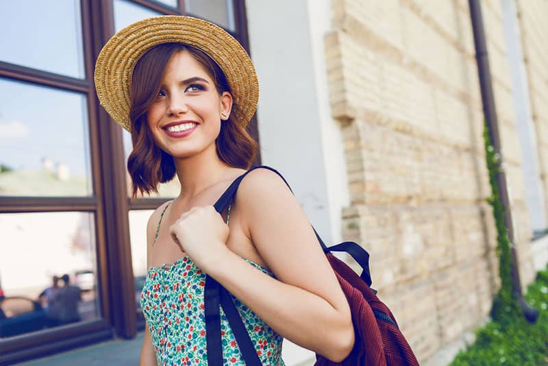 Ritratto di moda stile di vita soleggiato di giovane donna hipster alla moda che cammina per strada, indossando abiti alla moda, cappello di paglia, viaggio con zaino.