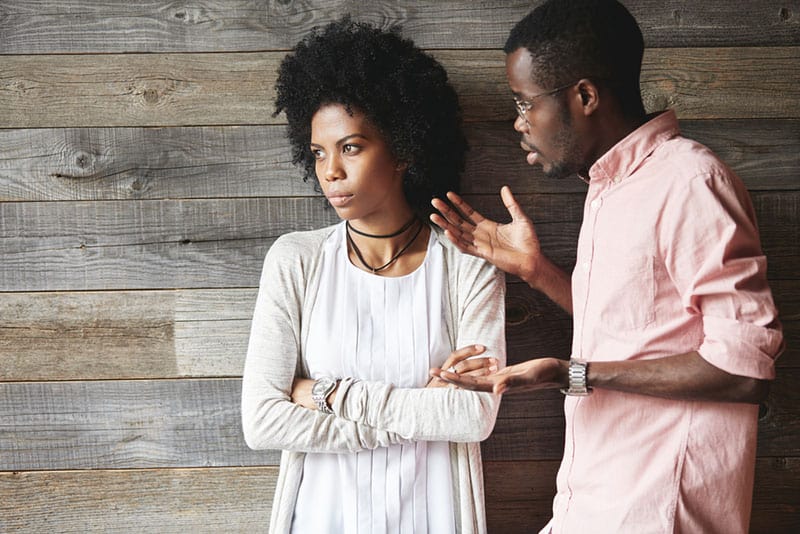 Young displeased black male gesturing in indignation, arguing with his stylish girlfriend, who is standing against wooden wall with folded arms, looking away with offended expression on her face