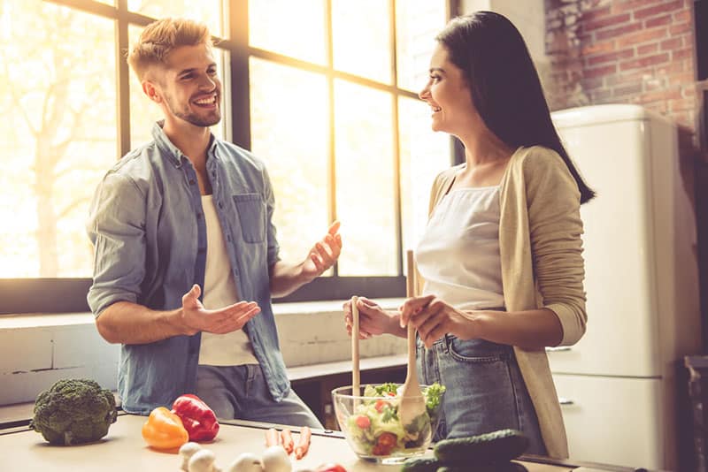 Hermosa pareja joven está hablando, mirando a la cámara y sonriendo mientras se cocina en la cocina en casa. La mujer está mezclando ensalada