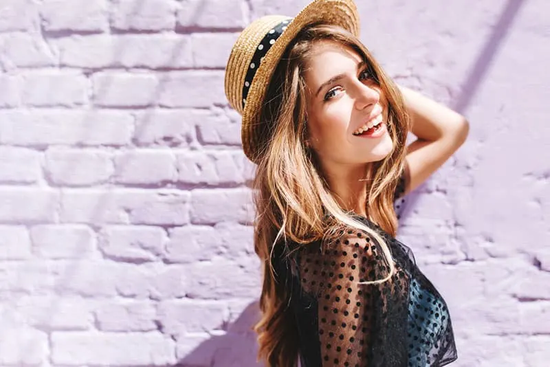 Graceful young woman with long shiny hair gladly posing while walk outside in good mood. Outdoor close-up photo of pleased girl in trendy summer hat with black ribbon enjoying sunshine.