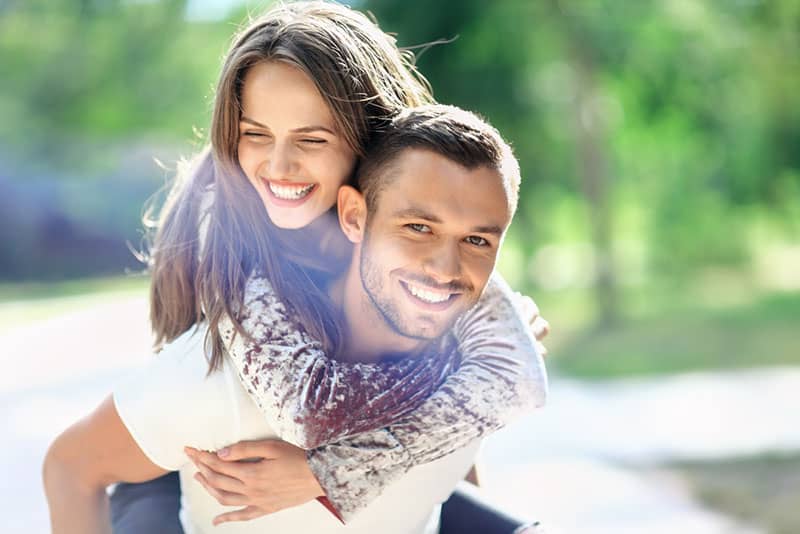 Man carrying his girlfriend on the back. Happy laughing couple piggyback. Young cheerful loving pair have fun together on sunny day. Love, romance, peoples relationship. Image with lens flare effect