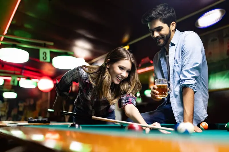 woman playing pool while smiling man looking at her with glass of beer