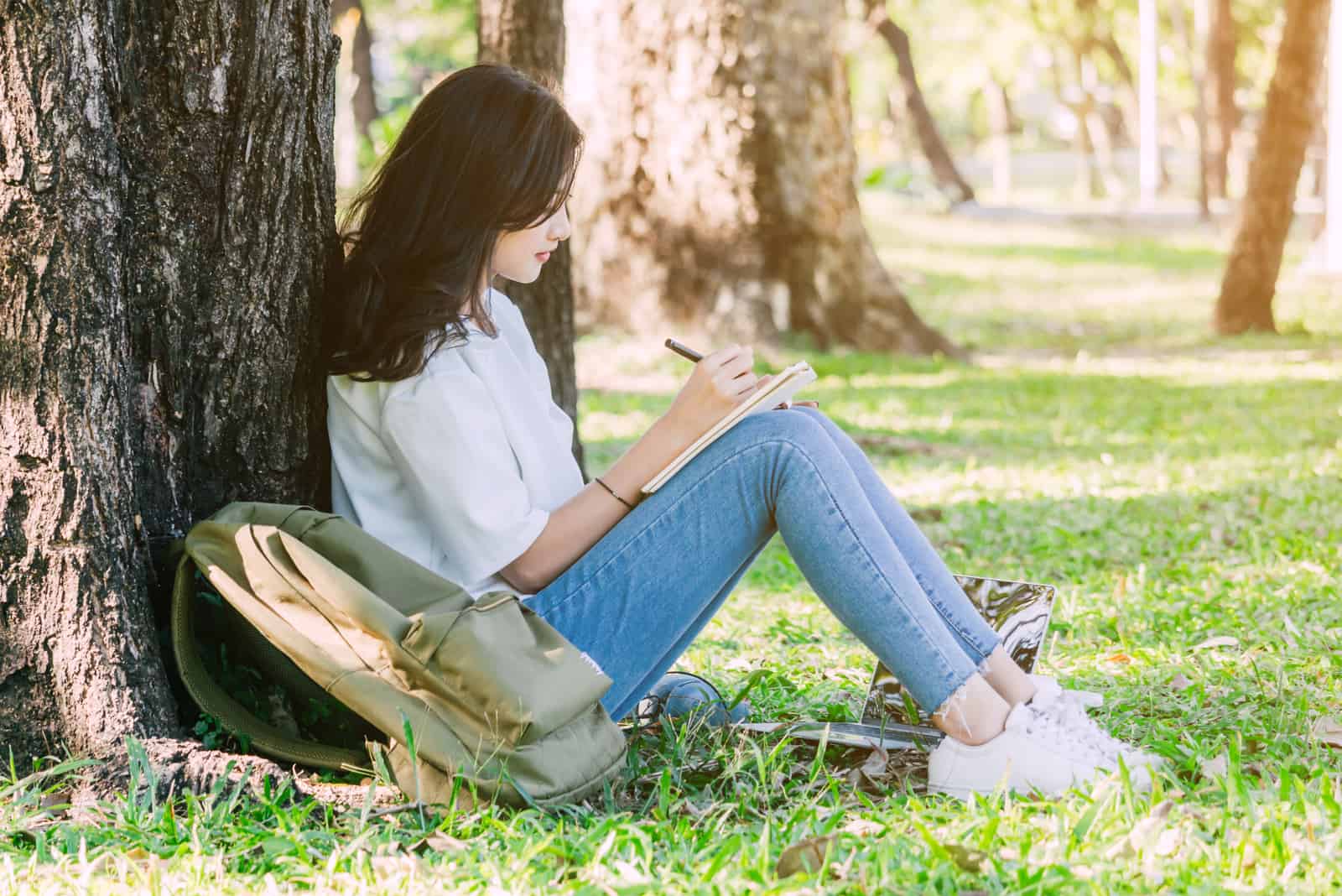a woman sits by a tree and writes a letter