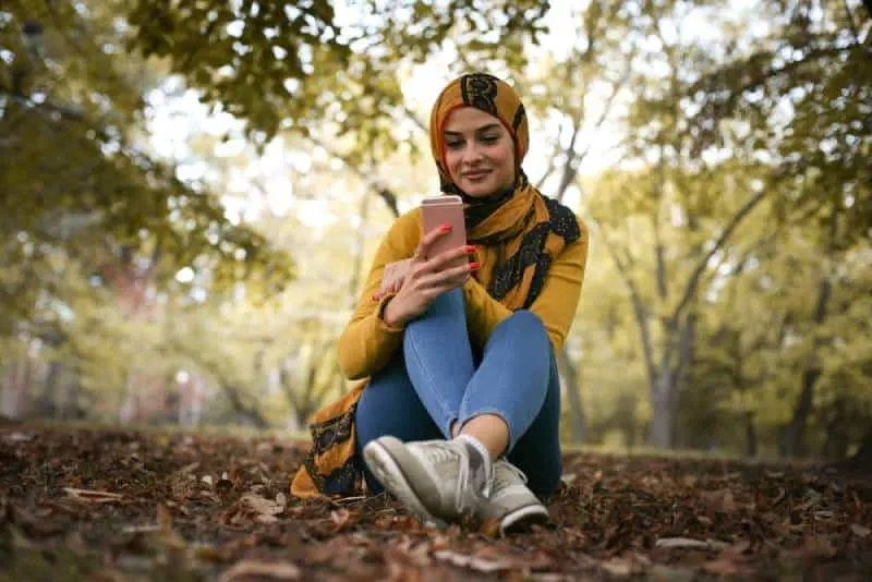 happy young woman sitting in forest and typing on phone