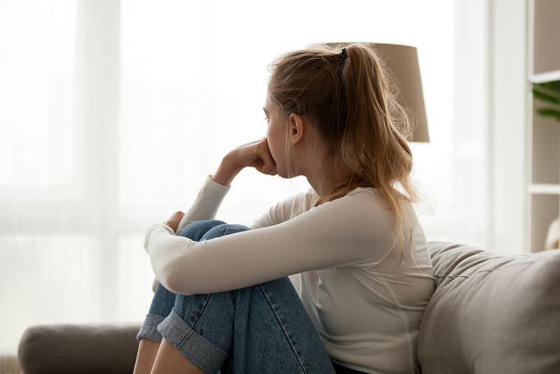 sad young blonde woman sitting on couch and looking through window