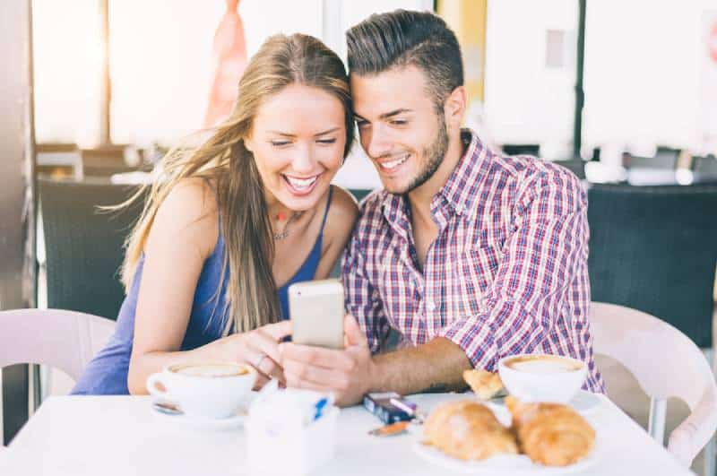 casal sorridente num café a tomar o pequeno-almoço e a olhar para o telemóvel