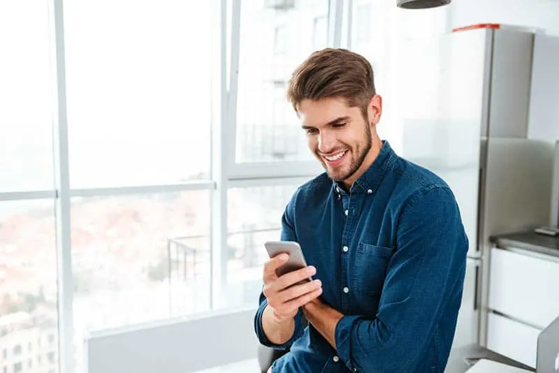 smiling man in blue shirt texting on his phone