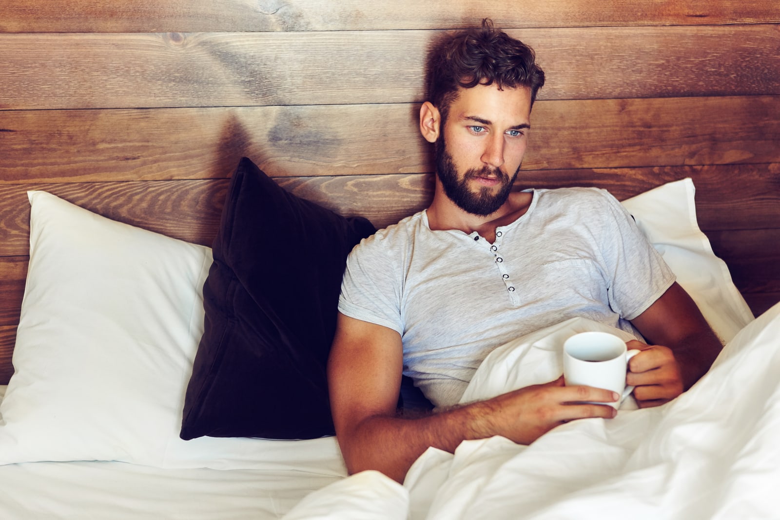 young man lying in bed and looking thoughtfully right while drinking morning coffee