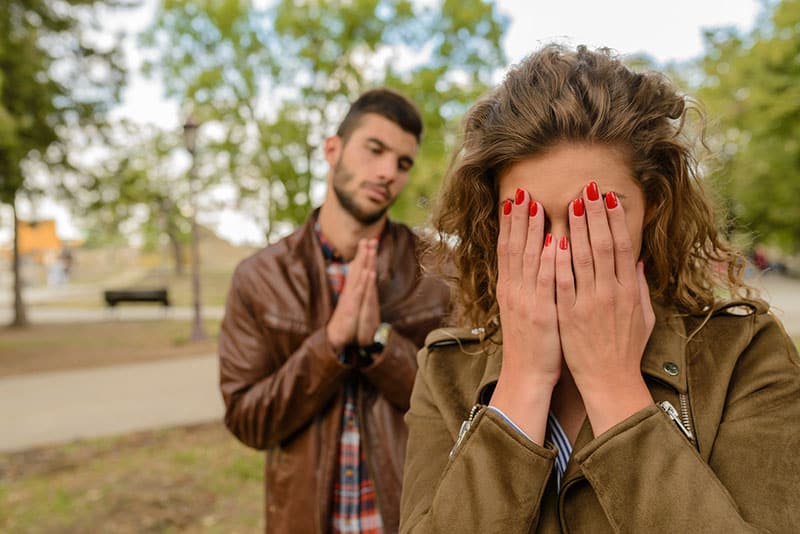 Donna piangente con giacca verde dietro l'uomo con giacca di pelle marrone che chiede perdono di giorno