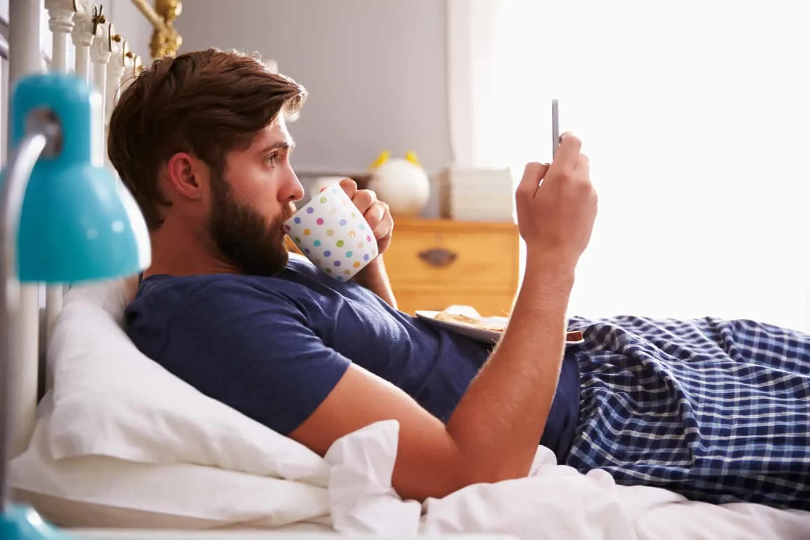 a man lies on the bed drinking coffee and pressing a phone