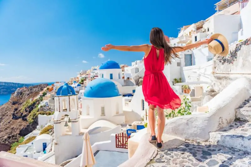 back view of traveling woman wearing red dress and standing with arms up in Santorini