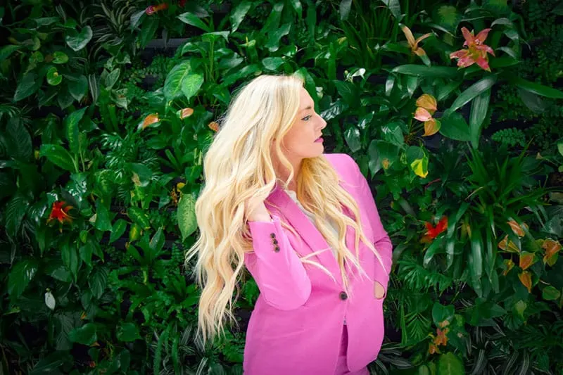 blonde girl in pink coat with green plants at the background
