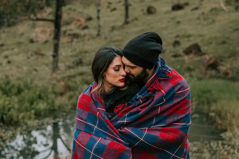 couple covered with blanket standing near a lake inside the woods