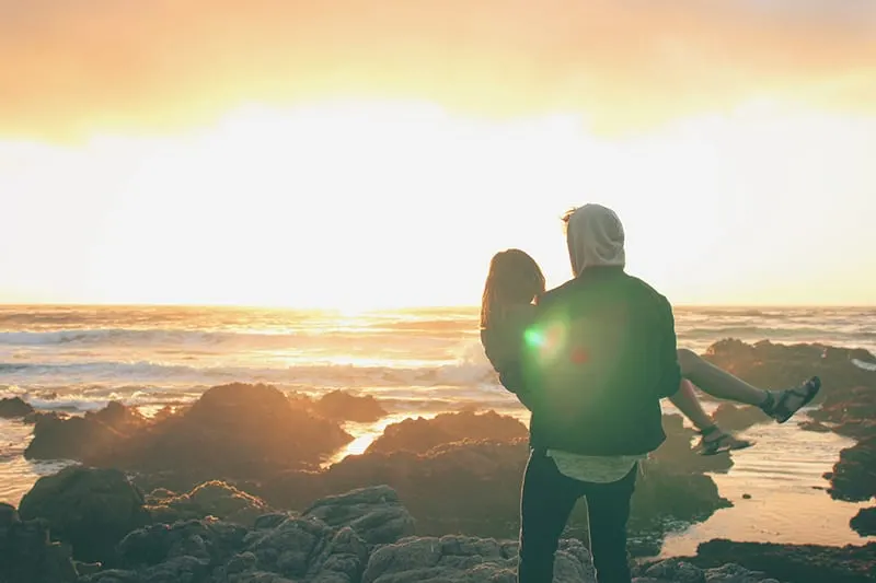 man carrying the woman on top of cliff near ocean during sunset