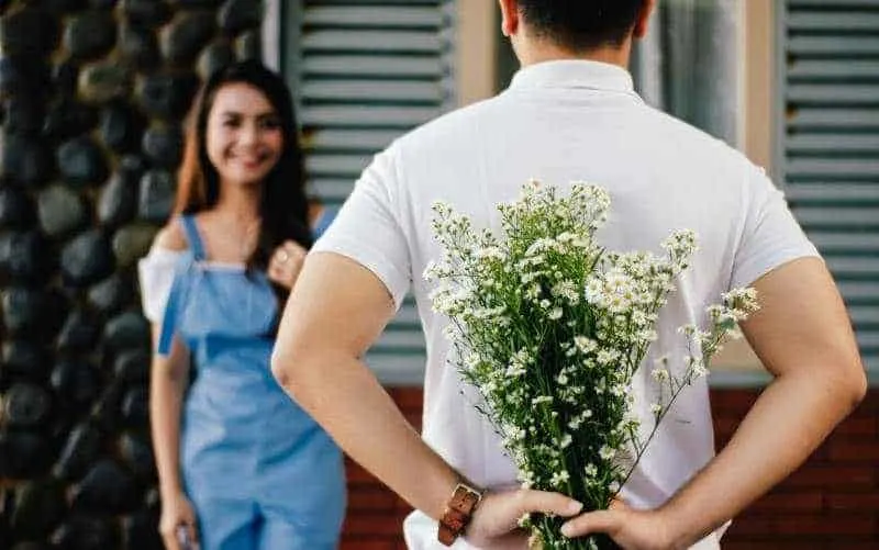 Man facing hapy woman with his hands on his back and flowers in his hands